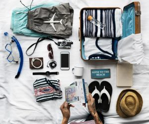 Flat lay of travel accessories including suitcase, clothing, camera, passport, phone, sandals, and beach items arranged on a bed.